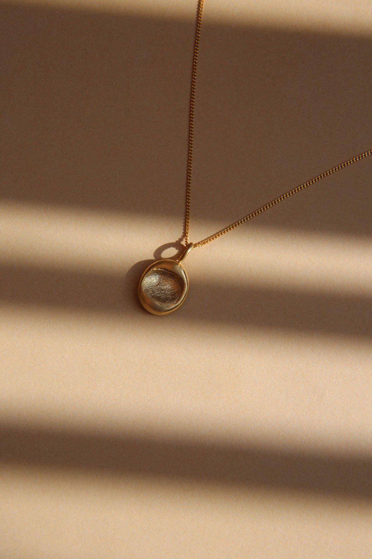 The details: small oval gold fingerprint pendant on a fine chain, in stripes of sunlight